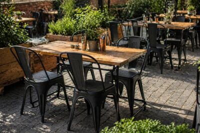 Outdoor cafe seating with wooden tables, black metal chairs, and potted plants in sunlight.|  Krzesło kawiarniane industrialne Metro PP – czarne | krzeslaonline.pl